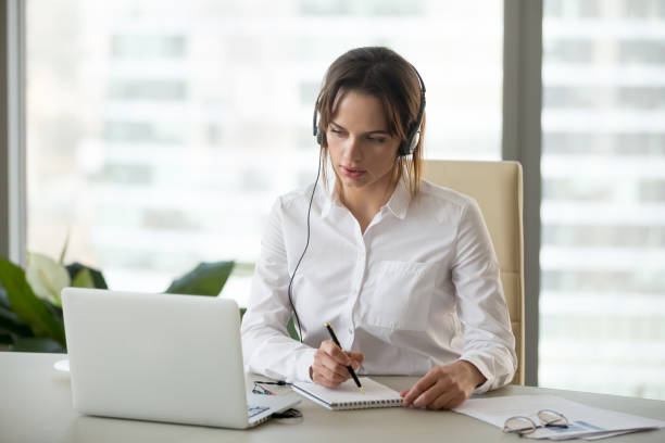 Serious businesswoman in headphones watching webinar on laptop making notes