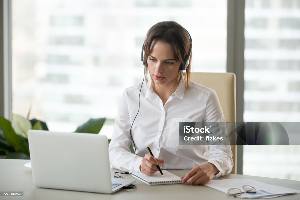 Mulher de negócios sério em fones de ouvido assistindo webinar no laptop, fazendo anotações - Foto de stock de Tradução royalty-free