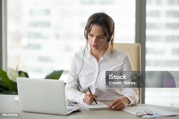 Foto de Mulher De Negócios Sério Em Fones De Ouvido Assistindo Webinar No Laptop Fazendo Anotações e mais fotos de stock de Tradução