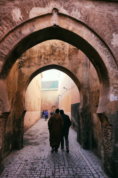 Marrakech, Morocco -January 18, 2018: Narrow red alley street in medina of Marrakech, Morocco. Silhouette of two men in traditional jellaba going through archways of old town on a sunny day.