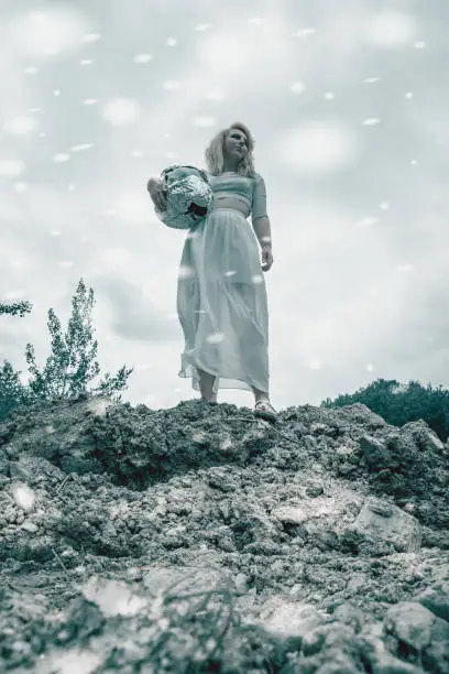 Low Angle Full Length View of Woman Wearing White Dress Standing Holding Helmet on Rocky Cliff Ledge and Looking into the Distance While Snow Falls Around Her in Fantasy Extraterrestrial Themed Image