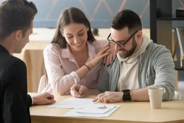 Excited millennial couple signing purchase agreement buying first home together, husband puts signature on document, becoming apartment owner, spouses legalize property ownership in Real Estate Agent office
