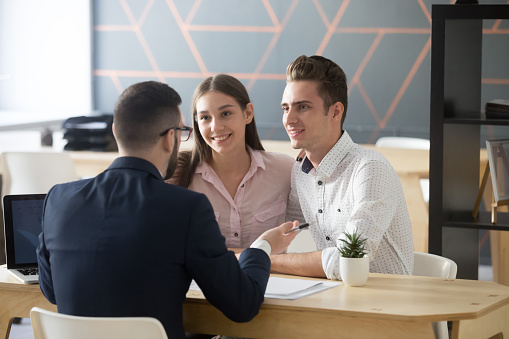 Insurance agent or broker consulting smiling millennial couple on property purchase, Real Estate Agent discussing mortgage loan with young spouses, explaining contract terms at meeting or negotiations in office