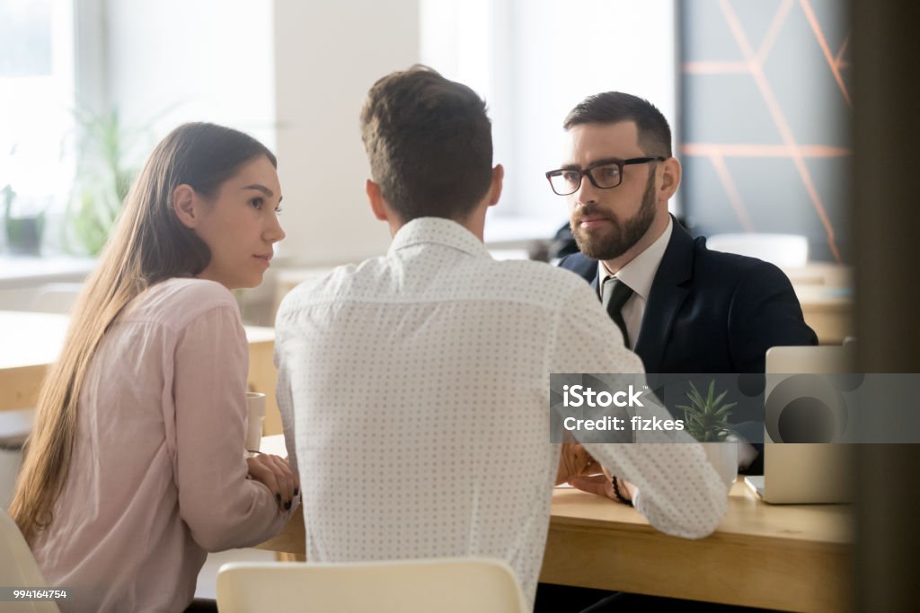 Broker listening to clients during office meeting or consultation Serious broker or Real Estate Agent listening to millennial couple arguments or ideas during office meeting, insurance agent consulting clients on house purchase, becoming property owners or taking loan. Lawyer Stock Photo