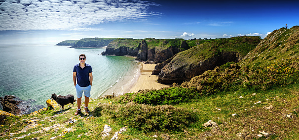 Single Hispanic man lifestyle portrait at beach standing on headland at Skrinkle Haven on the Pembrokeshire Coast with his dog