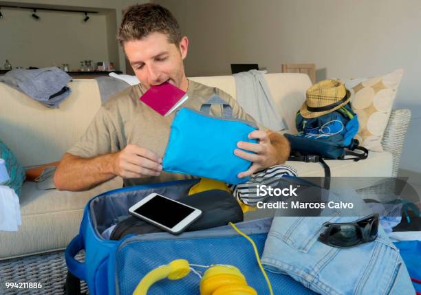 Young Attractive And Happy Man At Home Couch Preparing Travel Back And Packing Suitcase Folding Clothes And Organizing Passport And Things Before Leaving On Holidays Trip And Travel Concept Stock Photo - Download Image Now