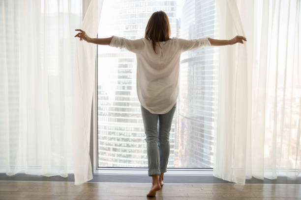 mujer de pie mirando por ventana abrir cortinas, vista trasera - high rise apartments fotografías e imágenes de stock