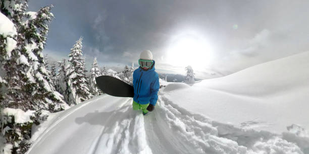 360 OVERCAPTURE 3D: Freerider walks uphill to start his descend down mountain covered in fresh snow. Male snowboarder walks up a narrow trail made in deep powder snow on a sunny day in the mountains.