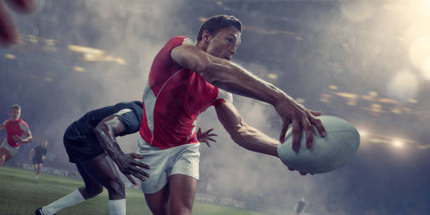 A male rugby player running and holding rugby ball in outstretched arms, about to throw the ball in a pass. The player wears a generic red and white strip, just about to be tackled by a rival player. The action takes place in a generic stadium under foggy, rainy conditions.