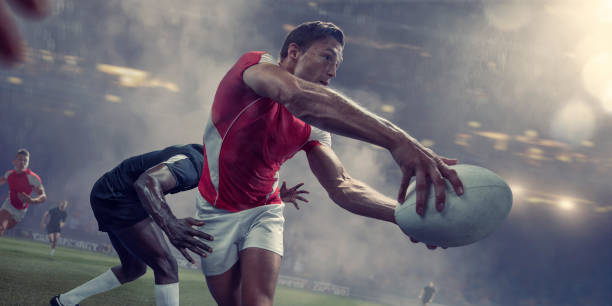 Rugby Player About To Pass Ball Just Before Being Tackled A male rugby player running and holding rugby ball in outstretched arms, about to throw the ball in a pass. The player wears a generic red and white strip, just about to be tackled by a rival player. The action takes place in a generic stadium under foggy, rainy conditions. rugby stock pictures, royalty-free photos & images