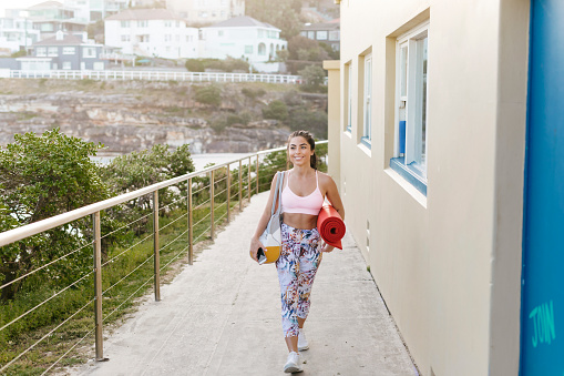 A beautiful woman in workout clothes is holding a smart phone in her hand. She is looking at the camera as she walks towards it. Carrying her workout equipment in a colourful duffel bag and a red exercise mat on her shoulder she is walking next to a houses.