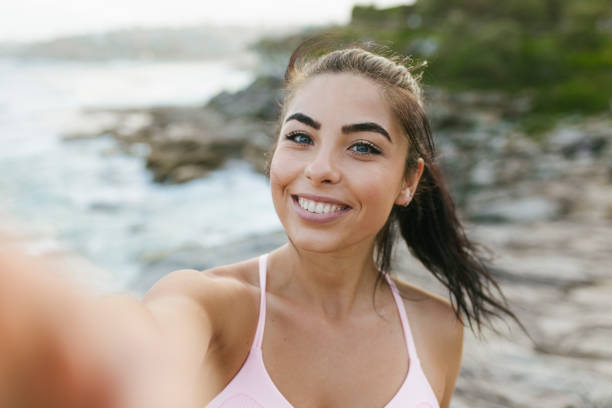 A portrait of a beautiful smiling woman holding a camera up in the air and looking straight into it, taking a selfie at a rocky seaside. Her hair blowing in the wind.