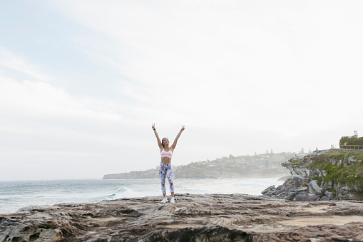 A beautiful and fit woman is cheering on a cliff overlooking the ocean. She is looking at the camera with her hands risen up in the air with her fingers making a peace sign.