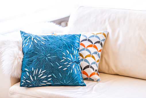 Closeup of two blue and orange pillows on couch or sofa by bright window in modern apartment, house or home with staging of large beige, neutral white colors