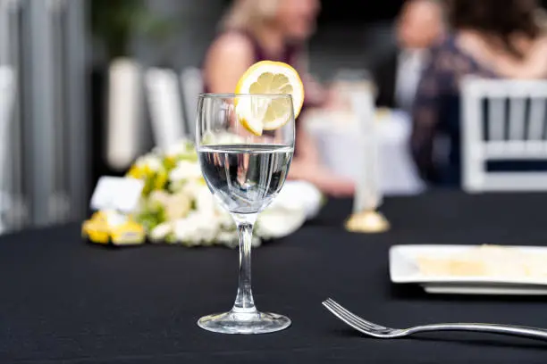 Closeup of one glass with lemon slice and water in restaurant or wedding reception black table tablecloth, bokeh of people, plate with cake slice, bouquet flowers, fork, chairs, dishes