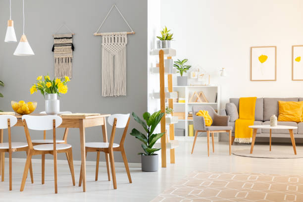 real photo of a spacious home interior with wooden table, white chairs and macrame on the wall in dining room and a gray sofa and armchair in the living room - wall flower sunny temperate flower imagens e fotografias de stock