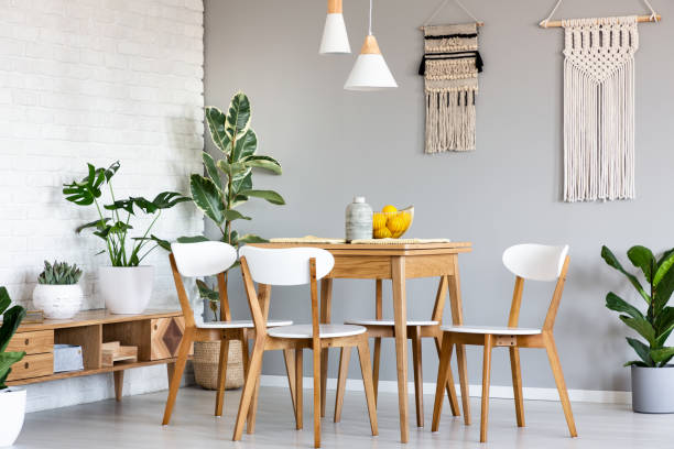 Macrame hanging on gray wall above wooden table and chairs in bright dining room interior with lots of plants. Real photo