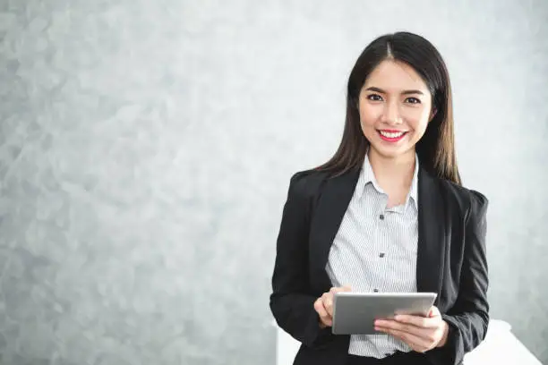 Photo of Portrait young Asian businesswoman holding tablet/smartphone in formal suit in office with copy space