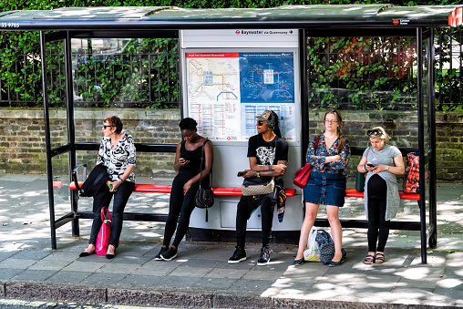 London, UK - June 22, 2018: People women waiting for bus at stop tired during day standing under cover, signs for Bayswater Queensway Underground tube metro on street road in city