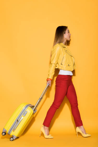 vista laterale di elegante viaggiatore asiatico femminile che trasporta borsa gommata su sfondo giallo - valigia a rotelle foto e immagini stock