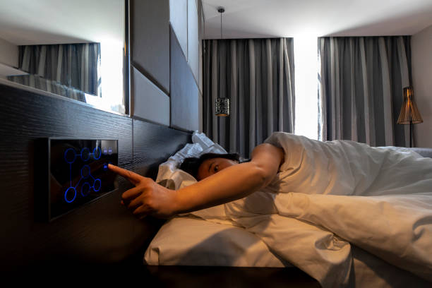 woman sleeping on bed, pushing button to open curtain, in a smart hotel stock photo