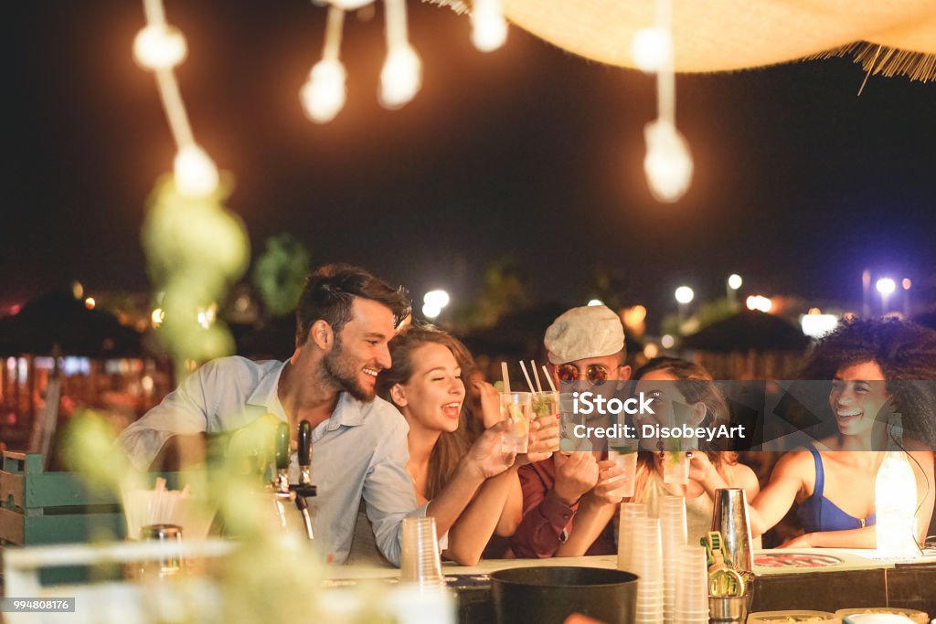 Happy friends cheering and drinking cocktails at beach party outdoor - Young millennials people having fun at weekend summer night - Youth lifestyle and nightlife concept - Main focus on left guys Beach Stock Photo