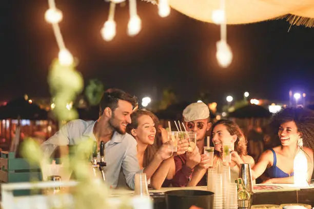 Photo of Happy friends cheering and drinking cocktails at beach party outdoor - Young millennials people having fun at weekend summer night - Youth lifestyle and nightlife concept - Main focus on left guys