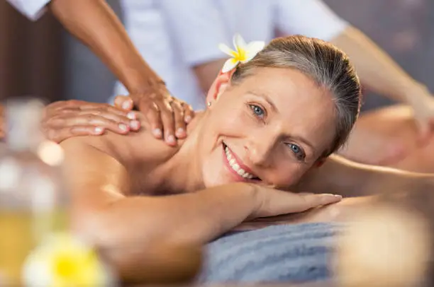 Happy senior woman getting oil massage on her back at spa center. Portrait of senior woman receiving back massage. Closeup face of smiling mature woman relaxing on massage table and looking at camera.