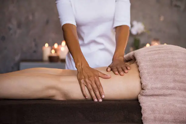 Therapeutic massage for senior woman by beautician at spa salon. Closeup of professional masseuse hands massaging woman legs at wellness center. Anti cellulite treatment and leg drainage on mature woman.