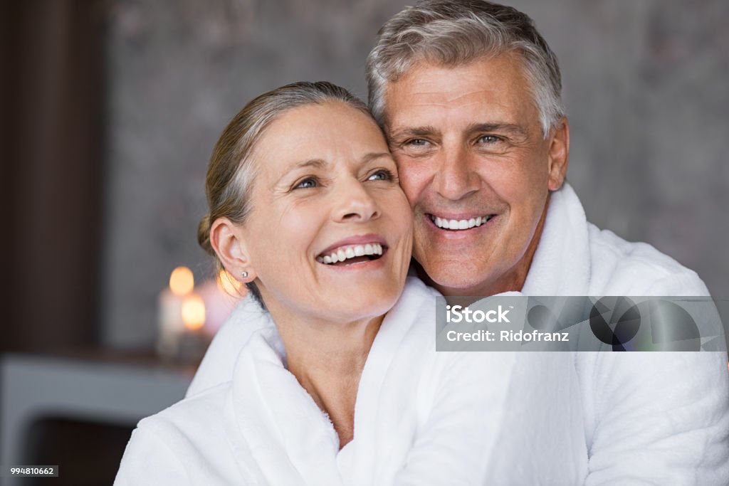 Laughing senior couple embracing at spa Smiling husband embracing cheerful wife from behind at spa. Laughing mature couple enjoying a romantic hug at wellness center after massage. Senior man and woman in white in bathrobe relaxing at spa. Spa Stock Photo