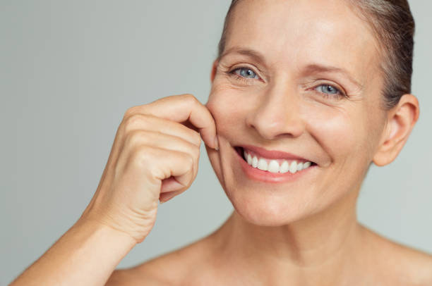 Beauty mature woman pulling perfect skin Senior woman pulling cheeks to feel softness and looking at camera. Beauty portrait of happy mature woman smiling with hands on cheek isolated over grey background. Aging process and perfect skin concept. cheek photos stock pictures, royalty-free photos & images