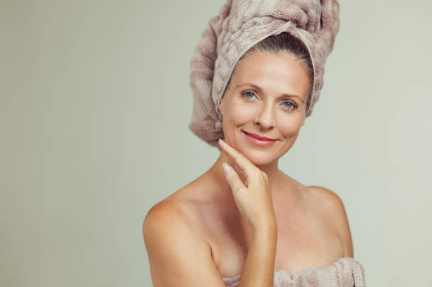 Beautiful mature woman in towel Portrait of smiling senior woman feeling soft skin with hand on chin after spa bath. Mature woman draped in towel on body and wet hair looking at camera. Lady in bathrobe after shower isolated on grey background. hair care women mature adult human skin stock pictures, royalty-free photos & images