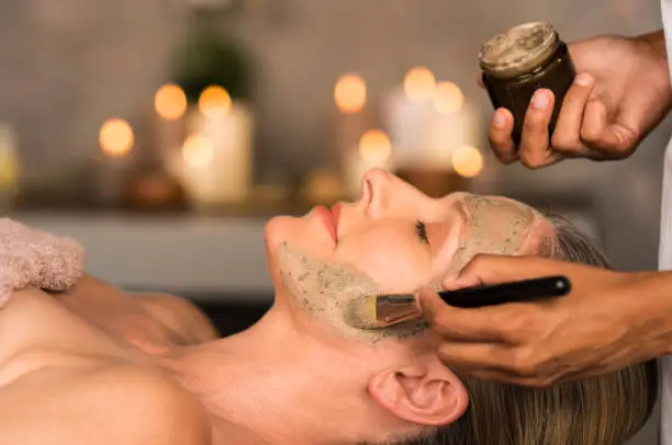 Senior woman relaxing while beautician getting purifying clay mask on her face. Closeup hand of masseuse applying mud from jar container on face of mature lady in spa center. Anti aging facial treatment with brush.