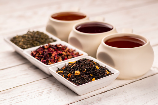 Various kind of tea in mugs and small square plates with dry tea on white wooden background