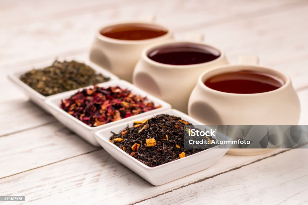 Three kind of tea in cups on wooden table Various kind of tea in mugs and small square plates with dry tea on white wooden background Tea - Hot Drink Stock Photo
