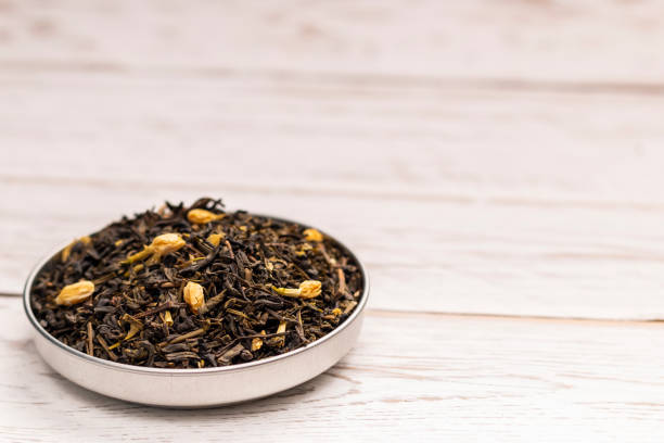 Close-up view of dry brown tea leaves with particles of jasmine flowers in small plate on white wooden table