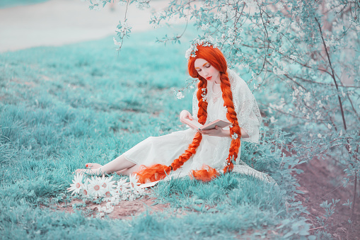 Young unusual redhead renaissance girl with curly hair braided in plait on a spring background. Beautiful woman with pale skin and in a renaissance dress reads a book in a meadow. Spring flowers