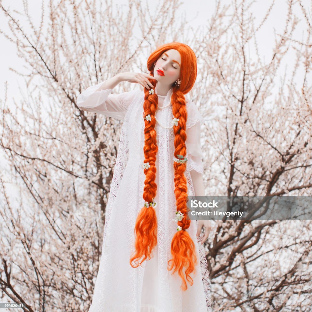 Young beautiful unusual redhead girl with curly hair braided in plait on a spring background. A woman with pale skin and very long red plait in a Pre-Raphaelite dress Redhead Stock Photo