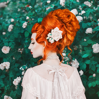 Beautiful redhead woman with high hairdo in a white dress on rose background. Portrait of young unusual pale girl with red hair. Beautiful model with stylish hairdo with a rose in garden