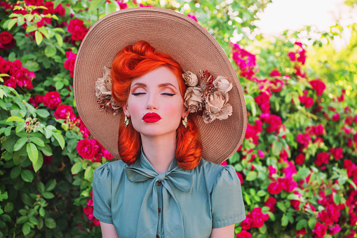 Luxury retro girl with red lips in mint dress on a beautiful flower summer background. Young redhead model in a hat with flowers on a background of a bush of roses. Stylish woman.