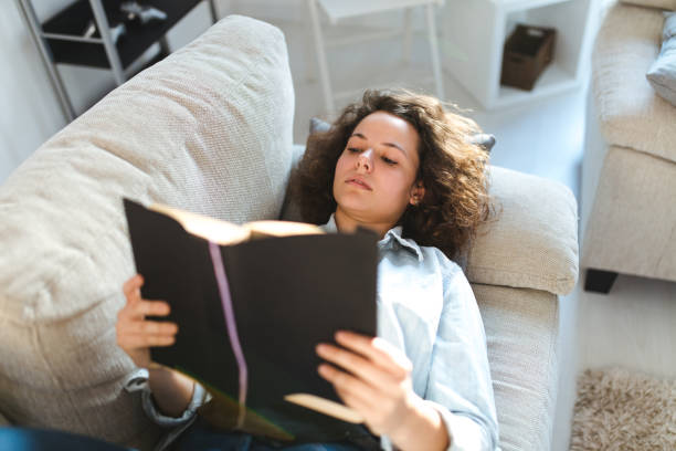 istock Happy young woman reading book on sofa 994823320
