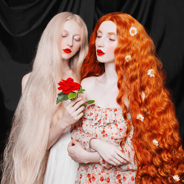 mujer joven hada con el pelo muy largo en vestido de flores sobre fondo negro. una hermosa chica con piel pálida. princesa de hadas renacentista con una rosa roja en las manos. dos hermanas. - peinados renacentistas fotografías e imágenes de stock