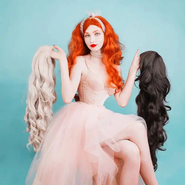 Photo of Hair care concept. Young beauty woman in long red wig, pale skin and in a pink dress on a blue background. Beautiful redhead model with long hair holds a white and black wig