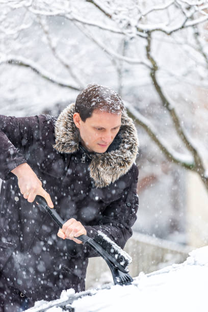 istock Young man cleaning car windshield from snow, ice with brush and scraper tool during snowfall while snowing snowflakes falling 994826888