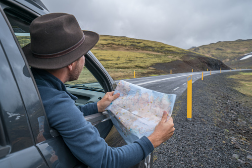 Cheerful caucasian male inside car looking at road map, green mountain and volcanic landscape. Road trip concept. \nTourist young man checking at tourist map.