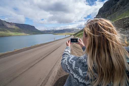 Cheerful caucasian female beside car takes mobile phone picture of the beautiful scenery green mountain and lake landscape. Road trip concept. \nBlond hair girl checking at tourist map.