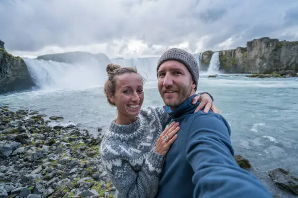 Travel couple fun taking selfie photo by Godafoss waterfall on Iceland using smartphone. People visiting famous tourist attractions and landmarks on Route 1