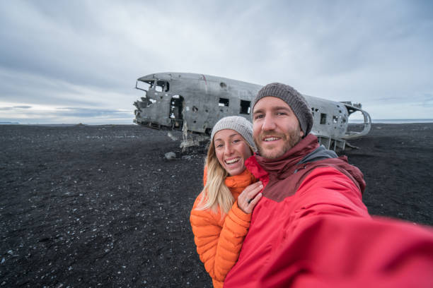 jovem casal em pé por acidente de avião na praia de areia preta, tomando um lugar famoso retrato de selfie para visitar na islândia e posar com o naufrágio - acidente evento relacionado com o transporte - fotografias e filmes do acervo