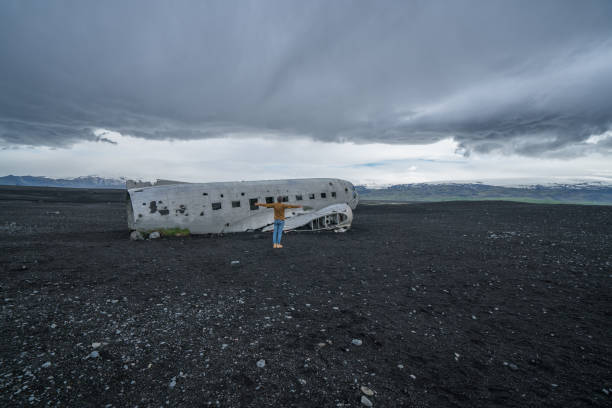 istock Young man stands arms outstretched near airplane crashed on black sand beach looking around her contemplating surroundings 994850424