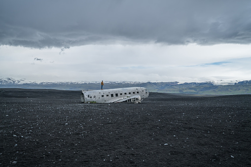 Young man stands on airplane crashed on black sand beach looking around her contemplating surroundings
Famous place to visit in Iceland and pose with the wreck